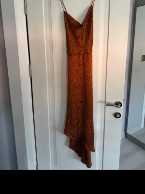 Lång brun prickig klänning med korsad rygg - Snygg långklänning i brun nyans med svarta prickar och tunna axelband. Klänningen är korsad i ryggen och ett mjukt, följsamt material som ger ett elegant fall. Endast använd 1 gång