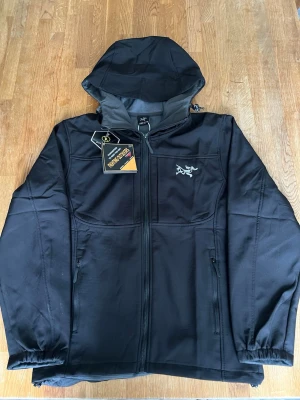 Arcteryx jacka  - Snygg svart vindjacka med huva och dragkedja framtill. Jackan har två sidofickor med dragkedja och ett diskret logomärke på bröstet. Materialet är slätt och vindtätt, perfekt för blåsiga dagar. Passar dig som vill ha en stilren och funktionell jacka. 