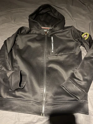 Svart hoodie Zenith Canada - Svart hoodie med dragkedja från Zenith Canada. Stor lönnlövsdetalj i grått på ryggen och patch på ena ärmen. Huva och två fickor framtill. Sportig och stilren design, perfekt för chill eller träning.