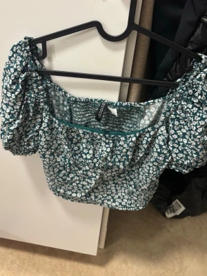 Grön blommig croppad topp  - Söt grön croppad topp med vitt blommönster. Puffiga korta ärmar och smockad rygg ger en trendig och bekväm passform. Perfekt till höga jeans eller kjol för en fräsch look.
