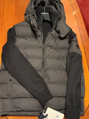 Svart hybrid jacka moncler - Snygg svart dunjacka med avtagbar huva och dragkedja framtill. Jackan har stickade ärmar och quiltad kropp för extra värme. Perfekt för kalla vinterdagar och har en modern, clean look.