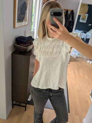 Vit topp med spets och volangärm - Supersöt vit topp med broderade spetsdetaljer och volangärmar. Hög hals och fina hålmönster ger en romantisk vibe. Perfekt till jeans eller kjol för en fräsch look. Luftigt material som passar perfekt till varmare dagar. Endast använd ett fåtal gånger 
