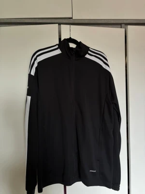 Svart Adidas träningsjacka - Säljer en svart träningsjacka från Adidas med vita ränder längs ärmarna och logga på båda ärmarna. Jackan har hel dragkedja. Perfekt för träning 