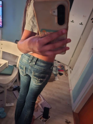 Blå bootcut jeans med slitningar - Säljer ett par blå bootcut jeans med slitningar på knäna och snygga broderade detaljer på bakfickorna. Jeansen har klassisk femficksdesign och är i stretchigt denimtyg. Perfekta för en avslappnad och trendig look.