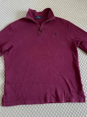 Vinröd halvzip från Polo Ralph Lauren - Säljer en vinröd långärmad tröja från Polo Ralph Lauren med halv dragkedja och hög krage. Tröjan har en liten blå logga broderad på bröstet och är tillverkad i mjukt bomullsmaterial. Perfekt för dig som gillar klassisk och stilren look.