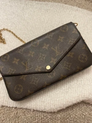 Louis Vuitton Felicie Pochette  - Säljer en brun clutch/väska från Louis Vuitton med klassiskt monogrammönster och guldfärgad kedja. Väskan har kuvertformad design och tryckknapp. Insidan är röd och har flera fack för kort och småsaker. Perfekt accessoar för dig som gillar lyxig stil.
