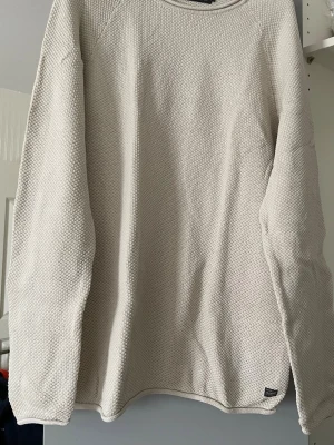 Beige strukturerad stickad tröja - En stilren beige stickad tröja med rund hals och lång ärm. Tröjan har en diskret struktur i materialet som ger en modern känsla. Passar perfekt till jeans eller kjol för en avslappnad look.