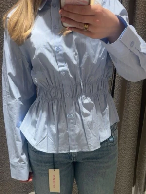 Blå randig blus med markerad midja - Snygg blå blus med tunna vita ränder och markerad midja med resår. Klassisk skjortkrage och knäppning framtill. Långa ärmar med breda manschetter ger en stilren look. Perfekt att matcha med jeans eller kjol.
