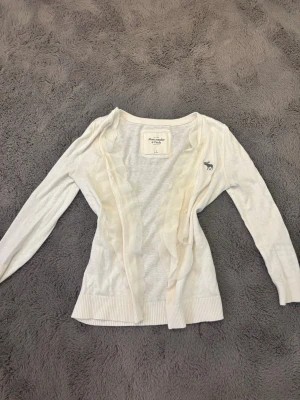 vintage abercrombie and fitch kofta - Säljer en tunn, vit kofta med volangdetaljer framtill och trekvartsärmar. Koftan är gjord i ett mjukt, tunt material som passar perfekt till lager på lager. Supersnygg att matcha med linne eller topp. super fin 2000s vibe, är lite nopprig på vissa ställen men inga stora defekter. inga knappar så en öppen kofta. köpte på sellpy. pris går att diskutera💕