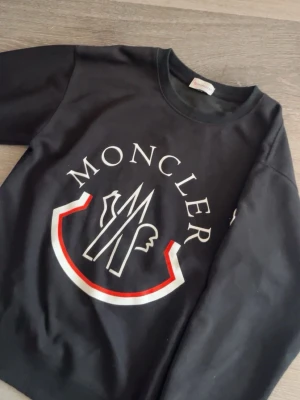 Svart Moncler sweatshirt med logga - Svart sweatshirt från Moncler med stor vit och röd logga framtill och klassisk rund hals. Tröjan har långa ärmar och en liten Moncler-patch på ena ärmen. Perfekt för dig som gillar streetwear och vill ha något stilrent men ändå iögonfallande.