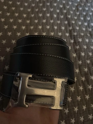 Svart bälte med silverspänne - Snyggt svart bälte med ett stort silverspänne i form av ett H. Bältet har vita sömmar längs kanterna och är gjort i skinn. Perfekt accessoar för att lyfta din outfit.
