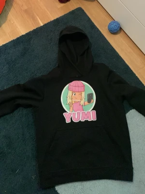 Svart hoodie med Yumi-tryck - Säljer en svart hoodie med stor färgglad Yumi-illustration på bröstet. Motivet visar en blond tjej i rosa mössa och tröja som tar en selfie, med texten 'YUMI' under. Hoodien har huva och magficka. Perfekt för dig som gillar snygga och unika prints.