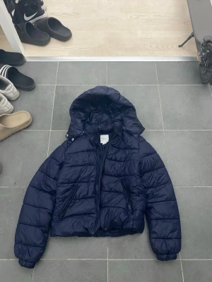 Mörkblå pufferjacka med huva - Snygg mörkblå pufferjacka med huva och dragkedja framtill. Jackan har två framfickor med dragkedja och quiltad design. Perfekt för kalla dagar och ger en trendig look. Materialet är glansigt och jackan har en klassisk pufferform. Är i strl 158