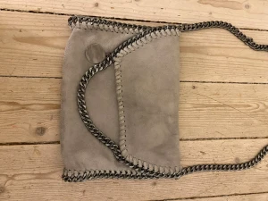 Grå axelväska - Snygg grå axelväska i mockaimitation med silverfärgade kedjor längs kanterna och som axelrem. Väskan har en vikbar front och dekorativ söm runtom. Perfekt för dig som gillar en trendig och stilren look.