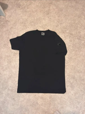 C.p 3ompany  - En svart t-shirt med korta ärmar och en unik rund detalj på bröstfickan. T-shirten har en enkel och clean design, perfekt för dig som gillar minimalistisk stil. Tillverkad i mjukt material för skön känsla hela dagen.