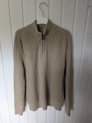 Beige ribbstickad tröja med half zip - Beige ribbstickad tröja med hög krage och half zip-dragkedja framtill. Tröjan har lång ärm och en relaxed passform, perfekt för kyligare dagar. Enkel och stilren design som passar till det mesta.