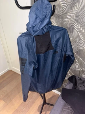 Blå vindjacka från The North Face - Snygg blå vindjacka från The North Face med huva och svart meshpanel på ryggen för extra ventilation. Stor logga på ärmen och mindre logga på bröstet. Perfekt för blåsiga dagar och streetwear-stil.