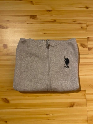 Grå hoodie från U.S. Polo Assn. - Snygg grå hoodie med dragkedja från U.S. Polo Assn. Tryckt logga på bröstet och detaljer på ärmen. Mjuk bomullsblandning, två fickor fram och justerbar huva med metallöljetter. Perfekt för en avslappnad och sportig stil. Använd 2-3 gånger. Pris kan diskuteras 
