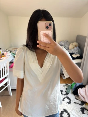 Vit v-ringad blus med puffärm - Snygg vit blus med v-ringning och puffiga korta ärmar. Dekorativa sömmar vid halsringning och ärmslut ger en stilren touch. Perfekt för dig som gillar en enkel men trendig look.