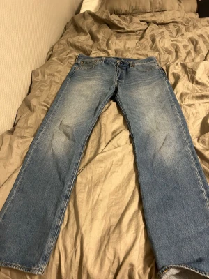 Blå Levis 501:or jeans - Klassiska blå jeans från Levi's med rak passform och fem fickor. Jeansen har en ljus tvätt med slitningar på låren och knäna för en avslappnad look. Tillverkade i denim, perfekt för dig som gillar tidlös stil. Knappt använt och pris kan diskuteras 