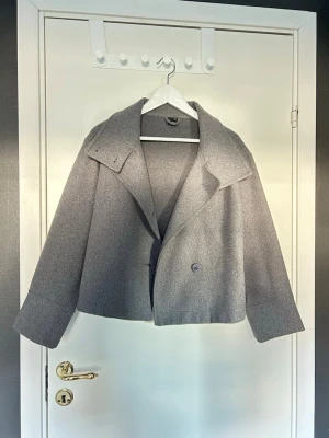 Grå kort ullkappa/ trenchcoat  - Stilren grå kappa i ullmix med bred krage och dubbelknäppning framtill. Modellen är kort och har raka ärmar samt en avslappnad passform. Perfekt för kyliga dagar när du vill ha en enkel men trendig look. Jätte nice är i storlek xxs men passar mig jättebra som vanligt bär storlek S. Aldrig använt är i jättte fint skick!!!