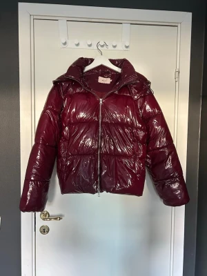 Vinröd glansig pufferjacka med huva - Snygg vinröd pufferjacka med glansig finish och stor huva. Jackan har dragkedja framtill och breda kanaler för extra värme. Perfekt för kalla vinterdagar och ger en trendig look. Materialet är syntetiskt och jackan har en boxig passform. Jätte varm och skön med bra fickor!!