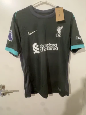 Liverpool bortatröja Konaté #5 Nike - Snygg mörkgrå Liverpool FC bortatröja med turkosa detaljer, tryck av Konaté och nummer 5 på ryggen. Nike-logga och Premier League-märke på ärmen. Tillverkad i lätt och ventilerande material, perfekt för match eller träning.