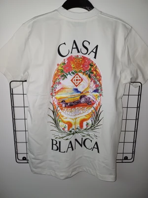 Vit t-shirt från Casablanca - Vit t-shirt från Casablanca med färgglatt tryck på både bröstet och ryggen. Trycket har blommor, bil och texten 'Casa Blanca'. Klassisk passform och rund halsringning. Perfekt för dig som gillar unika prints och stilrena plagg.