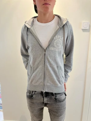 Grå zip-hoodie från Replay - Säljer en sjukt snygg grå zip-hoodie från Replay med tryck på bröstet och ryggen där det står 'Replay University'. Hoodien har dragsko i huvan, fickor fram och mjukt material. Perfekt för en chill och avslappnad stil. Modellen är 179 cm lång