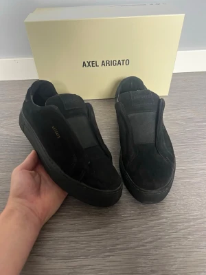 Svarta sneakers från Axel Arigato - Stilrena svarta sneakers från Axel Arigato i mocka med minimalistisk design och tjock sula. Skorna har diskret logga i guld på sidan och saknar synliga snören för en clean look. Perfekta för dig som gillar en modern och enkel stil.