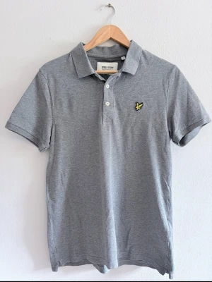 Grå piké från Lyle & Scott - Säljer en stilren grå pikétröja från Lyle & Scott med klassisk krage och knappslå. Tröjan har korta ärmar och den ikoniska gula loggan på bröstet. Tillverkad i mjuk bomull som känns skön mot huden.