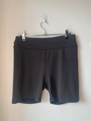 Svarta cykelshorts med bred midja - Säljer ett par svarta cykelshorts med bred midjeresår. De är stretchiga och har en enkel, clean look utan synliga detaljer eller mönster. Perfekta för träning eller chill dagar. Passformen är normal och materialet känns mjukt och följsamt.