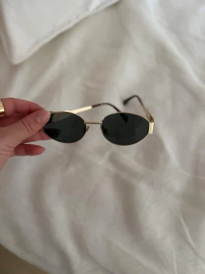 Ovala solglasögon med guldfärgade bågar - Snygga solglasögon med tunna, guldfärgade metallbågar och svarta ovala glas. Sidorna har en diskret logga i guld. Perfekta för att lyfta din stil under soliga dagar.