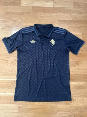 Juventus Mörkblå Fotbollströja - Mörkblå pikétröja från Adidas i samarbete med Juventus. Tröjan har diskret mönster i tyget, guldfärgade Adidas-logga och Juventus-emblem på bröstet samt guldfärgad 'JUVENTUS' text i nacken. Klassisk krage och korta ärmar. Perfekt för fotbollsfans.