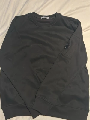 Svart CP Company sweatshirt - En stilren svart CP company sweatshirt med rund halsringning och ribbade muddar vid ärmslut och nederkant. Tröjan har en diskret svart patch på ena ärmen som detalj. Perfekt basplagg för en avslappnad look.