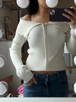 Offshoulder ribbad vit topp med dragkedja - Trendig offshoulder topp i ribbad vit trikå med lång ärm och dragkedja framtill. Snygg croppad passform som passar perfekt till jeans. Materialet känns mjukt och stretchigt.