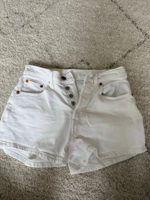 Vita högmidjade jeansshorts Levi's - Säljer ett par vita högmidjade jeansshorts från Levi's med knappgylf och klassiska fem fickor. Shortsen har en relaxed passform och är perfekta för varma dagar. Levi's-lappen syns tydligt bak i midjan och de har en tidlös look.💕