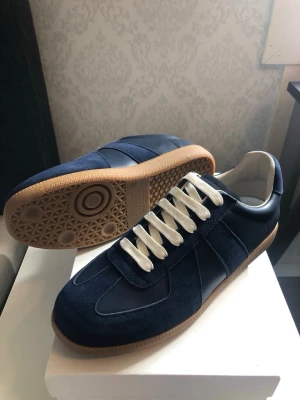 Maison Margiela  - Snygga marinblå sneakers med ovandel i mocka och släta paneler, vita snören och klassisk gummisula i ljusbrunt. Skorna har en stilren och tidlös design som passar till det mesta. Perfekta för dig som gillar en clean look.