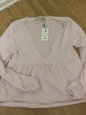 Ljusrosa v-ringad blus med spets - Supersöt ljusrosa blus med djup v-ringning och spetsdetaljer längs urringningen. Långärmad modell med lätt rynkad midja för en feminin siluett. Perfekt att styla med jeans eller kjol för en trendig look.