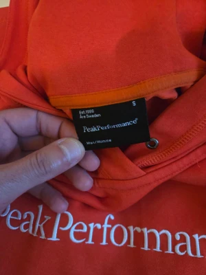  hoodie Peak Performance S - Säljer en orange hoodie från Peak Performance i storlek S. Hoodien har en klassisk passform med huva och vit broderad logga på bröstet. Perfekt för dig som vill ha en stilren och bekväm tröja till vardags. Materialet känns mjukt och skönt.