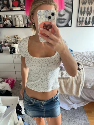 Vit blommig croppad topp - Söt vit croppad topp med småblommigt mönster i blått och grönt. Toppen har korta ärmar och fyrkantig urringning, vilket ger en trendig och feminin look. Perfekt att matcha med jeans eller kjol för en fräsch stil.