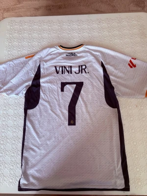 Real Madrid matchtröja Vini Jr #7 - Säljer en vit Real Madrid matchtröja från Adidas med Vini Jr och nummer 7 på ryggen. Tröjan har svarta och orange detaljer, klubbmärke på bröstet och sponsortryck. Tillverkad i lätt och ventilerande material, perfekt för fotbollsträning eller match.