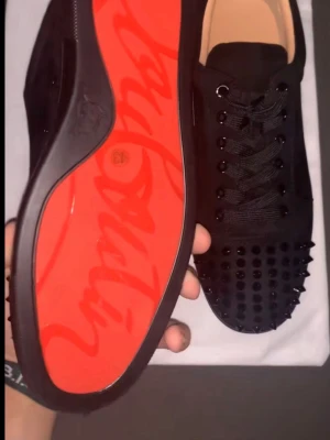 Svarta sneakers med nitar och röd sula - Säljer ett par svarta sneakers från Christian Louboutin med ikonisk röd sula och svarta nitar på tån. Skorna har snörning, ovandel i mocka och detaljer i svart läder. Insidan är beige och märkt med guldtext. Perfekt för dig som vill sticka ut.