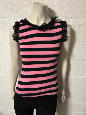 Topp med spets från Poizen S/M - Säljer en snygg ärmlös topp med rosa och svarta ränder. Toppen har spetsdetaljer vid axlar och halsringning samt en liten rosett framtill. Perfekt för dig som gillar en edgy och lekfull stil. Helt ny. Stretchig bomullstrikå. Storlek S/M.