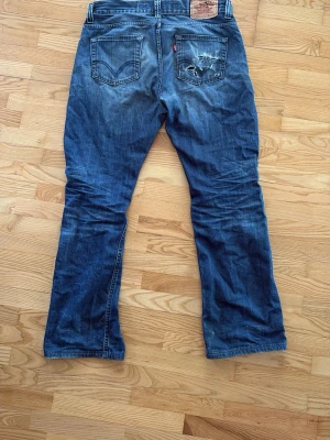 Vintage Levis 506 - Klassiska blå jeans från Levi's med raka ben och femficksmodell. Jeansen har slitningar och en patch bak i midjan. Materialet är denim i bomull och färgen är mellanblå med ljusare partier. Perfekta för en avslappnad och tidlös stil.