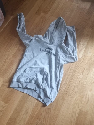 Grå hoodie från Peak Performance - Snygg grå hoodie från Peak Performance med svart logga på bröstet. Klassisk känguruficka framtill och stor huva. Tillverkad i mjukt bomullsmaterial, perfekt för chill dagar eller när du vill ha en avslappnad stil.