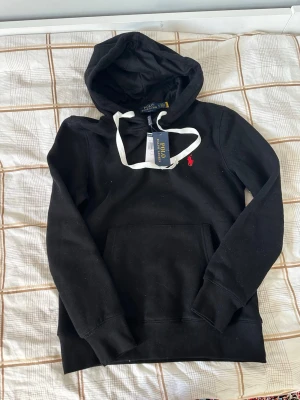Svart hoodie från Polo Ralph Lauren - Säljer en svart hoodie från Polo Ralph Lauren med vit dragsko och klassisk röd logga på bröstet. Hoodien har känguruficka framtill och ribbade muddar vid ärmar och nederkant. Perfekt för en chill och stilren look.