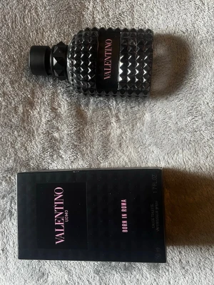 Valentino Uomo Born In Roma parfym - Säljer en Valentino Uomo Born In Roma parfym. Flaskan är svart med nitar och har en lyxig, modern design. Märket står tydligt på flaskan och kartongen, båda i svart med rosa text. Perfekt för dig som gillar exklusiva dofter och snygg inredningsdetalj.