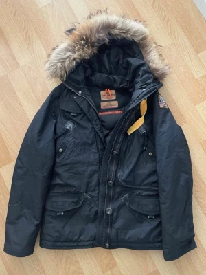 Svart Parajumpers parkas med pälskrage - Säljer en svart Parajumpers parkas från Masterpiece Series med avtagbar huva och pälskant. Jackan har flera fickor, gul detalj vid halsen och patch på ärmen. Perfekt för kalla dagar och riktigt snygg streetstil.Dragkejda är sönder och behöver repareras.