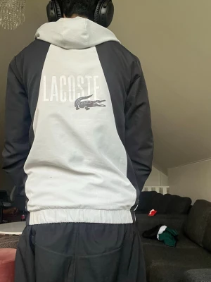 Lacoste svartvit vindjacka med huva - Snygg svartvit vindjacka från Lacoste med dragkedja framtill och huva. Jackan har svarta ärmar, vit kropp och stor Lacoste-logga med krokodil på ryggen. Elastisk midja och muddar för skön passform. Perfekt för en sportig och avslappnad stil.
